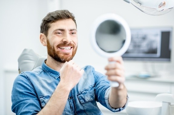 Man in denim shirt admiring his smile in mirror after dental treatment